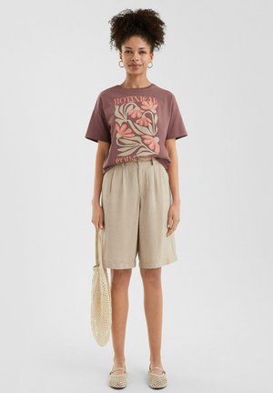 Brown graphic t-shirt with floral design, beige linen shorts, beige slip-on shoes, and a netted shoulder bag.