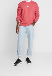 Rotes Sweatshirt mit weißem Nike-Logo, hellblaue Jeans mit hochgekrempelten Säumen und schwarze Sportschuhe mit weißen Akzenten. Einfarbiger Hintergrund.