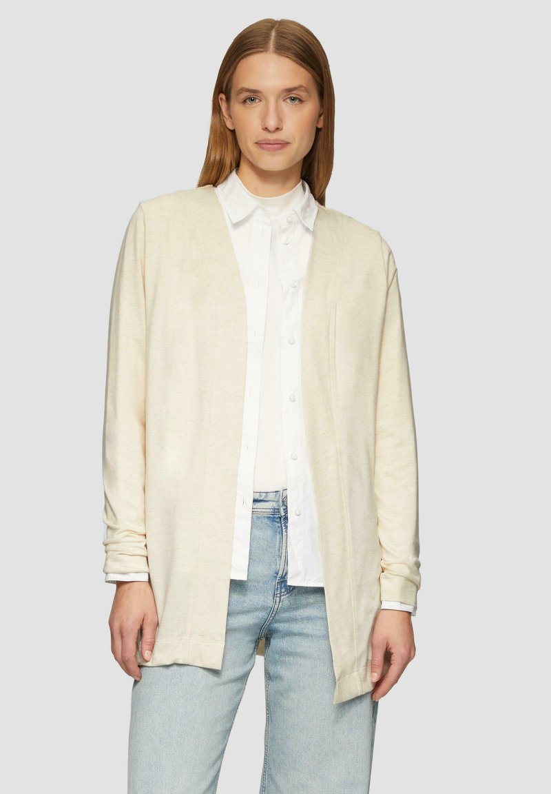 Lichtbeige cardigan gemaakt van zacht materiaal. Open ontwerp met lange mouwen, met een relaxte pasvorm en zonder knopen. G gedragen over een wit shirt.