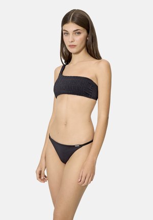 Mujer joven con un bikini negro texturizado de un solo hombro y braga bikini a la cadera a juego, de pie frente a un fondo claro y liso.