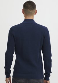 Mornarsko pleteno pulover s teksturiranim, vafljastim vzorcem. Ima visok ovratnik, rebraste manšete in rob. Čista zasnova, brez vidnih logotipov ali poudarkov.