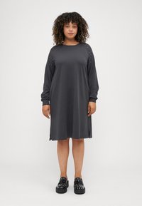 Grijze jurk met lange mouwen en zijsplitsen, met studaccenten op de schouders. Glad materiaal, relaxte pasvorm, knie-lange ontwerp. Zwarte platform schoenen.