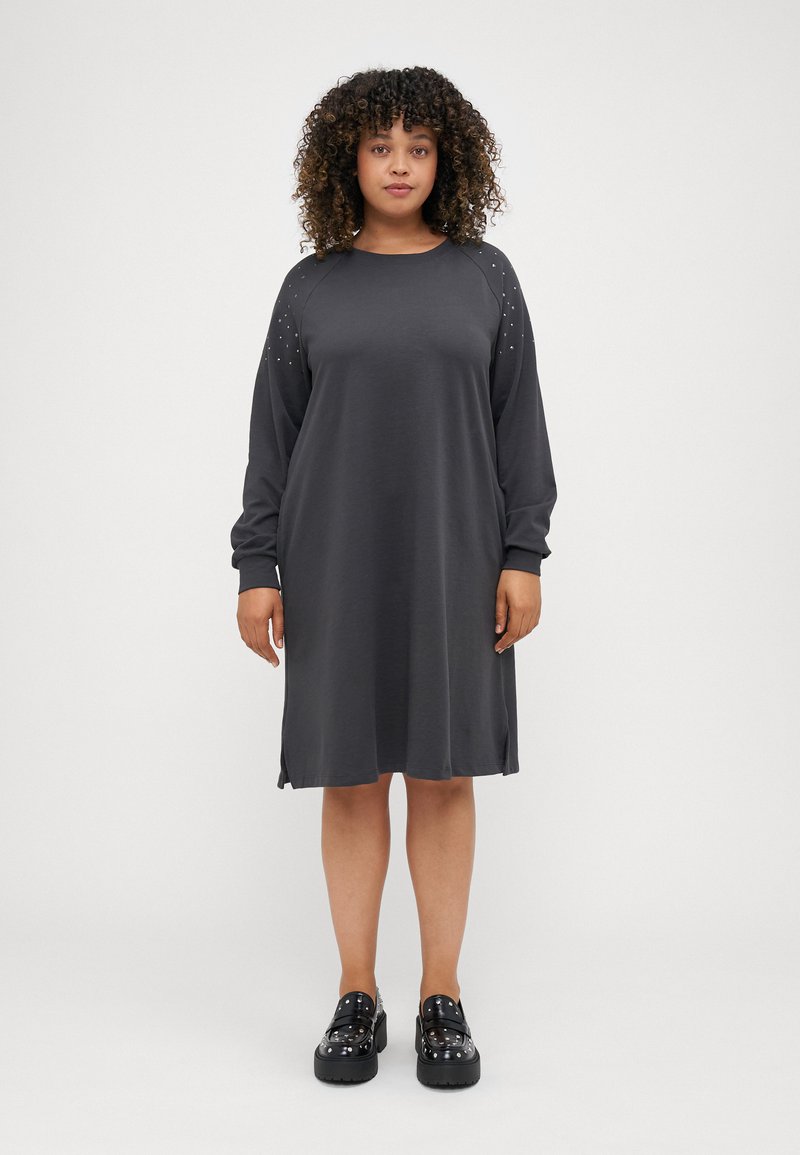 Grijze jurk met lange mouwen en zijsplitsen, met studaccenten op de schouders. Glad materiaal, relaxte pasvorm, knie-lange ontwerp. Zwarte platform schoenen.