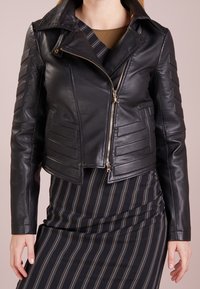 Musta nahkabiletakki, jossa on lyhyt malli, kulmikkaat paneelit ja kultaketju. Yhdistetty raidoitettuun mekkoon alla.