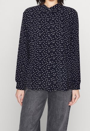 Frau trägt eine langärmelige, marineblaue Bluse mit weißen Punkten und einem runden Kragen, kombiniert mit locker sitzenden grauen Jeans.