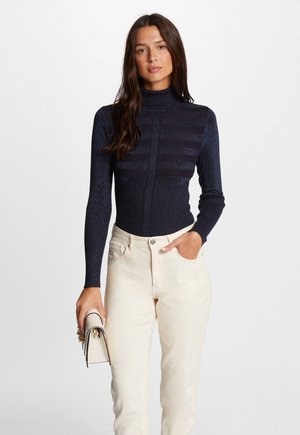 Marinblå långärmad polotröja med subtil glans och randig textur, tillsammans med ljusa högmidjade jeans och en vit handväska.