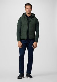 Gilet trapuntato verde con cappuccio sopra una maglietta scura, abbinato a pantaloni blu navy e sneaker nere. Presenta tasche con zip e dettagli con logo.