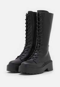 Black knee-high boots featuring a smooth leather material, prominent lacing, a side zipper, and a thick, textured platform sole.