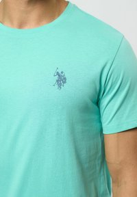 Ljus turkos t-shirt i mjuk bomull, med en liten grafisk tryck av en polo-spelare på en häst i mörkblått, rundhalsad design.