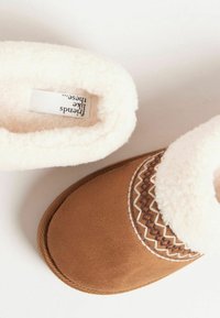 Barna velúr papucs fehér szőrme béléssel, díszítő geometrikus mintázattal a felső szélen. Simább tapintású, zárt kialakítású.