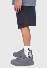 Graue Sneakers mit strukturierter Oberfläche und gepolstertem Kragen, kombiniert mit dunklen Shorts mit einem dezenten Logo und weißen Socken mit schwarzer Schrift.
