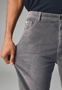 Szare spodnie sztruksowe z fakturą prążkowaną, przednimi kieszeniami i dopasowanym krojem. Zapięcie na guziki i suwak w talii zapewniające pewne dopasowanie.
