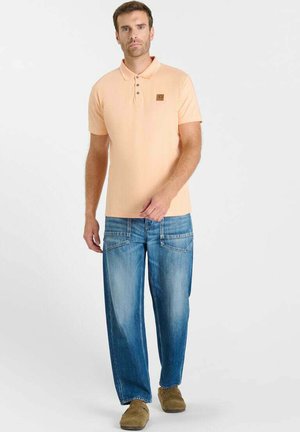 Man die een perzikkleurig poloshirt met korte mouwen draagt, blauwe spijkerbroek met voorzakken en bruine instapschoenen, staand tegen een effen achtergrond.