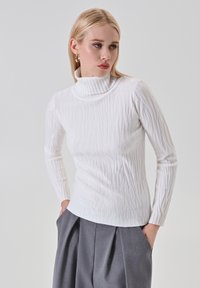 Donna bionda che indossa un maglione a collo alto bianco con texture e pantaloni grigi plissettati, con le mani in tasca, mentre guarda di lato.