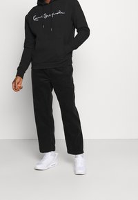 Czarna bluza z kapturem z białym logo w stylu skryptu, zestawiona z czarnymi spodniami z korduroy i białymi trampkami. Prosty design o luźnym kroju.