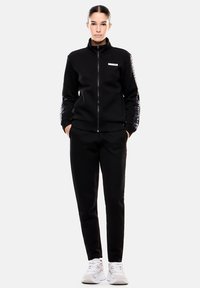 Chaqueta negra con cremallera y cuello alto, con mangas texturizadas, combinada con pantalones a juego. Presenta un logo blanco en la chaqueta.