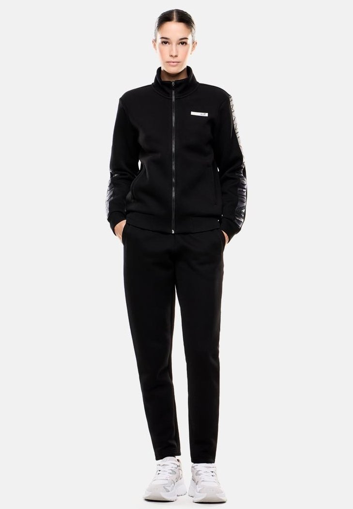 Chaqueta negra con cremallera y cuello alto, con mangas texturizadas, combinada con pantalones a juego. Presenta un logo blanco en la chaqueta.
