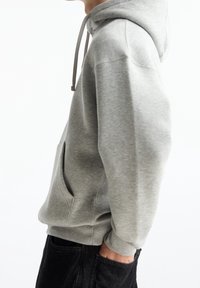 Grijze hoodie van zacht materiaal, voorzien van een voorvak, capuchon met trekkoord en geribbelde manchetten. Gestyled met zwarte broek.