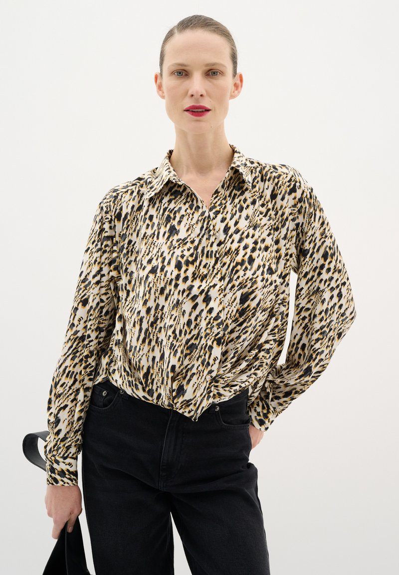 Blusa de manga larga con estampado de leopardo en tonos de crema, negro y marrón. Cuenta con un cuello y detalle anudado en la cintura, combinada con pantalones negros.