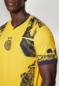 Kollane jalgpallijersey, millel on V-kaela disain ja mustad graafilised mustrid õlgadel ja rinnal. Logo ja tähed on selgelt nähtavad.