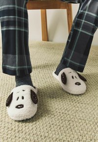 Bílé chlupaté pantofle s hnědým obličejem a ušima psa; nošeny přes tmavě šedé ponožky. Vzorované kárované kalhoty jsou viditelné na texturovaném koberci.