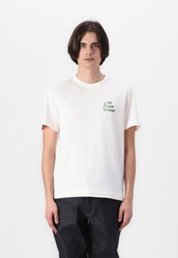Hvid bomulds t-shirt med korte ærmer. Har et grønt grafisk print på venstre bryst med teksten "THE CLUB SCENE." Enkel design, rund hals.