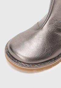 Nærbilde av metallisk sølv lær støveltå med detaljert søm og lysebrunt gummisåle mot hvit bakgrunn.