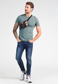 Conjunto casual que incluye una camiseta verde ajustada, jeans oscuros y una riñonera de camuflaje. Zapatillas con una mezcla de gris y negro.