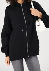 Svart zip-up huvtröja med avslappnad passform, frontfickor och dragsnören. Matchad med ljusgrå jeans och en liten vit handväska.