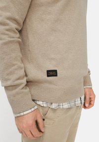 Beige knitted sweater featuring ribbed cuffs and hem, with a black label on the side stating "MADE FOR DAILY ADVENTURES." Layered over a plaid shirt.