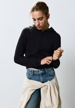 Kvinde iført en sort hættetrøje med lynlås og blå jeans med en beige jakke bundet omkring taljen, kigger ned og justerer sin ærme.