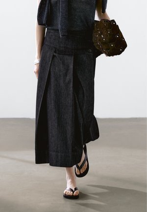 Personne portant une longue jupe noire, un haut noir, des sandales noires, tenant un sac à main marron texturé et un bracelet argenté au poignet gauche.