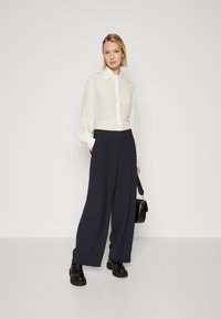 Camicia bianca con maniche lunghe e leggermente voluminose abbinata a pantaloni a gamba larga blu scuro a vita alta e stivali neri massicci, mentre tiene una borsa nera.