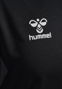 Schwarzes Sportoberteil mit einem auffälligen weißen Hummel-Logo, das ein stilisiertes Bienenmotiv über dem Markennamen in einer klaren Schriftart zeigt.
