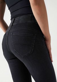 Kvinna som bär svarta högmidjade skinnyjeans med bakfickor och en svart åtsittande topp, stående mot en enkel ljus bakgrund.