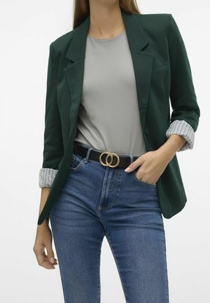 Femme portant un blazer vert foncé avec des poignets rayés roulés, un haut gris clair, un jean bleu et une ceinture noire avec une boucle double anneau dorée.