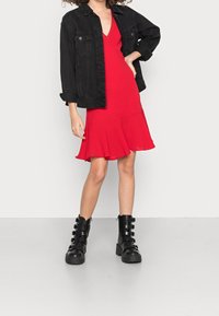Vestido rojo sin mangas con un dobladillo acampanado, complementado con una chaqueta de mezclilla negra y botas negras gruesas con múltiples correas y un tacón grueso.