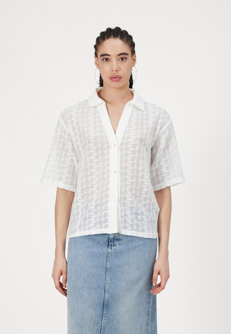 Camicia bianca a maniche corte con motivo testurizzato e colletto aperto, dotata di chiusura a bottoni e vestibilità comoda, abbinata a una gonna in denim.