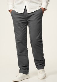 Gray chinos made of soft fabric, featuring a straight cut. Paired with white sneakers, held hands in pockets, and a white shirt.