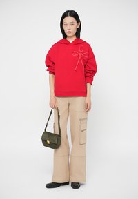 Camisola com capuz vermelha com um detalhe decorativo de laço, combinada com calças cargo bege. Acessorizada com uma pequena mala verde e sapatos pretos.