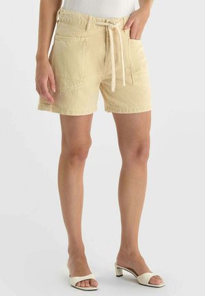 Femme portant un short beige à cordon avec des poches avant et des sandales blanches à talons ouverts, debout devant un fond uni.