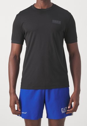 Mann trägt ein schlichtes schwarzes T-Shirt mit kurzen Ärmeln und leuchtend blaue Sportshorts mit weißen Logos am linken Bein.