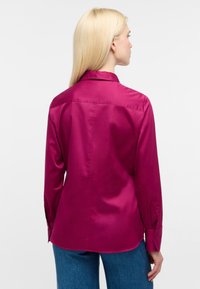 Blusa di raso magenta con colletto, maniche lunghe, polsini con bottoni e orlo curvo. Texture liscia con un riflesso lucido.