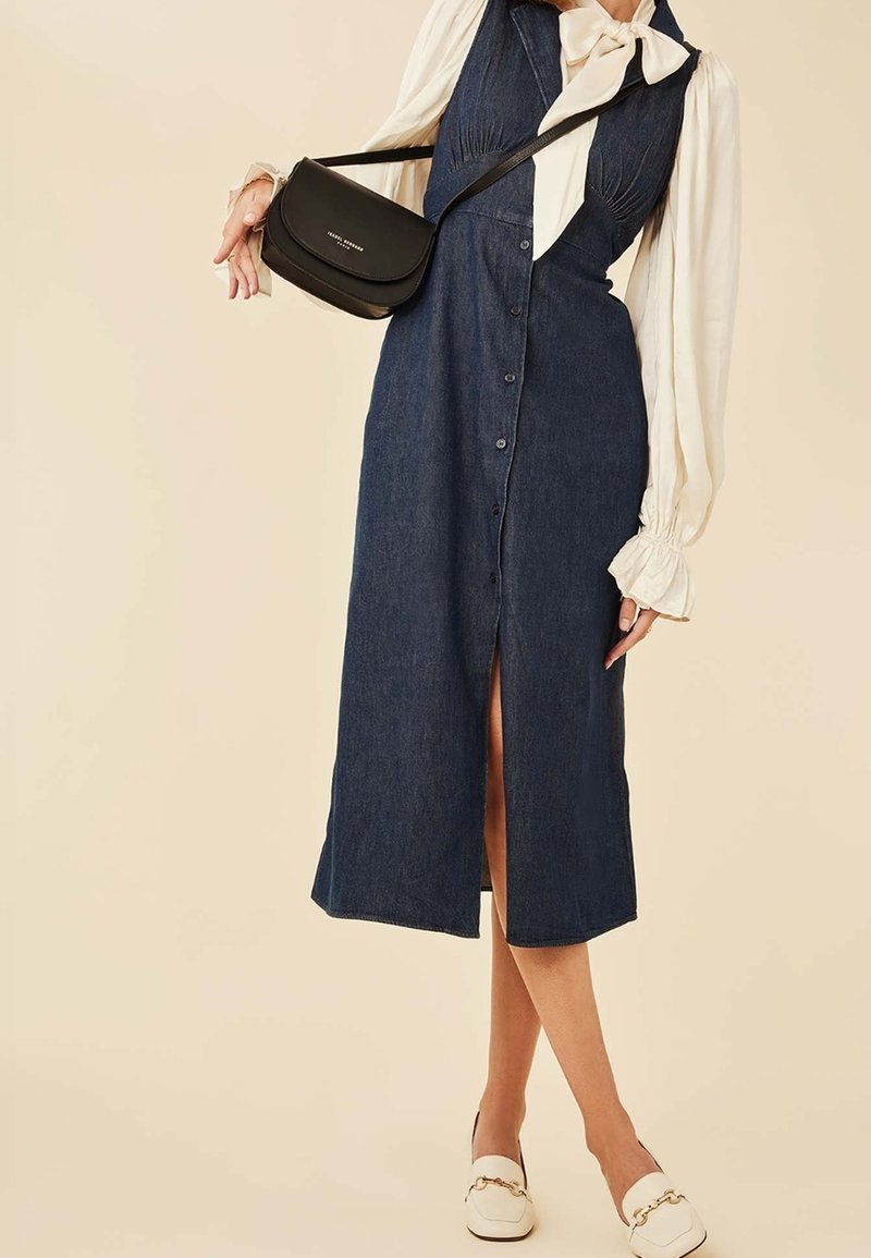 Denim midi-jurk met voorbouten, een split en een aansluitend lijfje. Gecombineerd met een zwarte schoudertas en witte instapschoenen. Lichte achtergrond.
