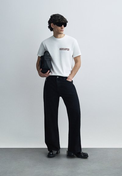 Άσπρο βαμβακερό μπλουζάκι με το κείμενο "KIDSUPER", μαύρο τζιν παντελόνι με φαρδιά πόδια, κρατώντας μια μικρή μαύρη τσάντα, φοράει μαύρα γυαλιά ηλίου και παπούτσια πλατφόρμα.