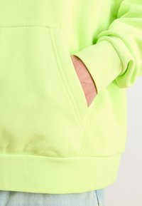 Neongrön huvtröja med ficka fram, ribbade ärmar och en texturerad tyg. Visar en avslappnad design med en rak nederkant.