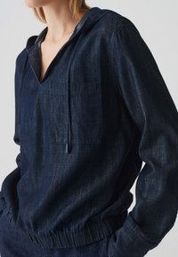 Felpa con cappuccio in denim blu navy, dotata di tasca frontale, orlo elastico e coulisse. Tessuto testurizzato con una leggera lucentezza.