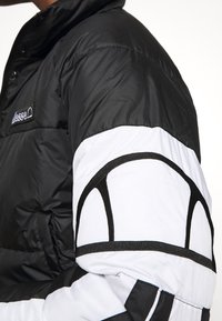 Manche d'une veste matelassée noire et blanche avec un design en demi-cercle noir et un petit écusson logo rectangulaire sur la poitrine.