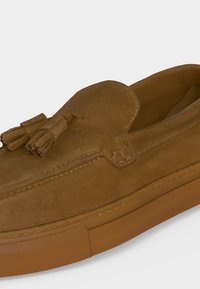 Καφέ μοκασίνι από σουέτ με λεπτομέρεια με φούντες και λαστιχένια σόλα, κοντινή λήψη σε ουδέτερο φόντο.