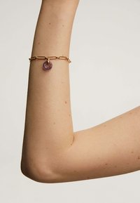 Pulsera de cadena de oro con eslabones alargados y un charm redondo y translúcido en color púrpura. La pulsera reposa sobre un brazo de tono de piel claro.